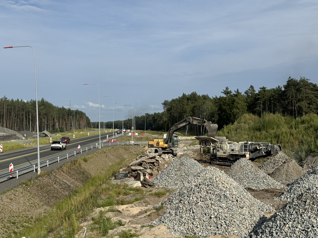 Construction site with excavator and gravel piles next to a highway bordered by forest.