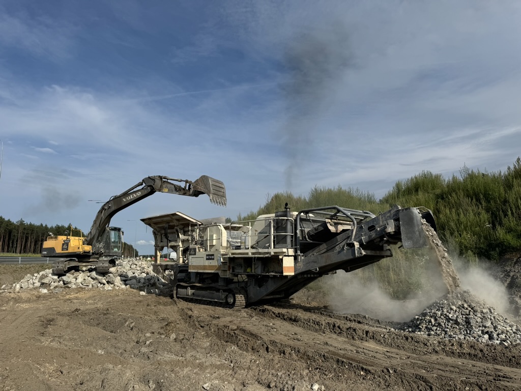 Excavator loading rocks into crusher machine that deposits crushed stones into piles outdoors.