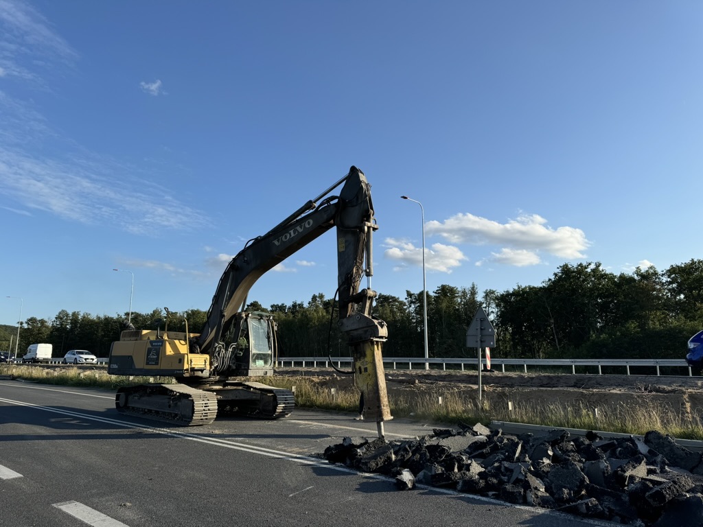 Volvo excavator breaking up asphalt road on a sunny day with blue sky and trees in background.