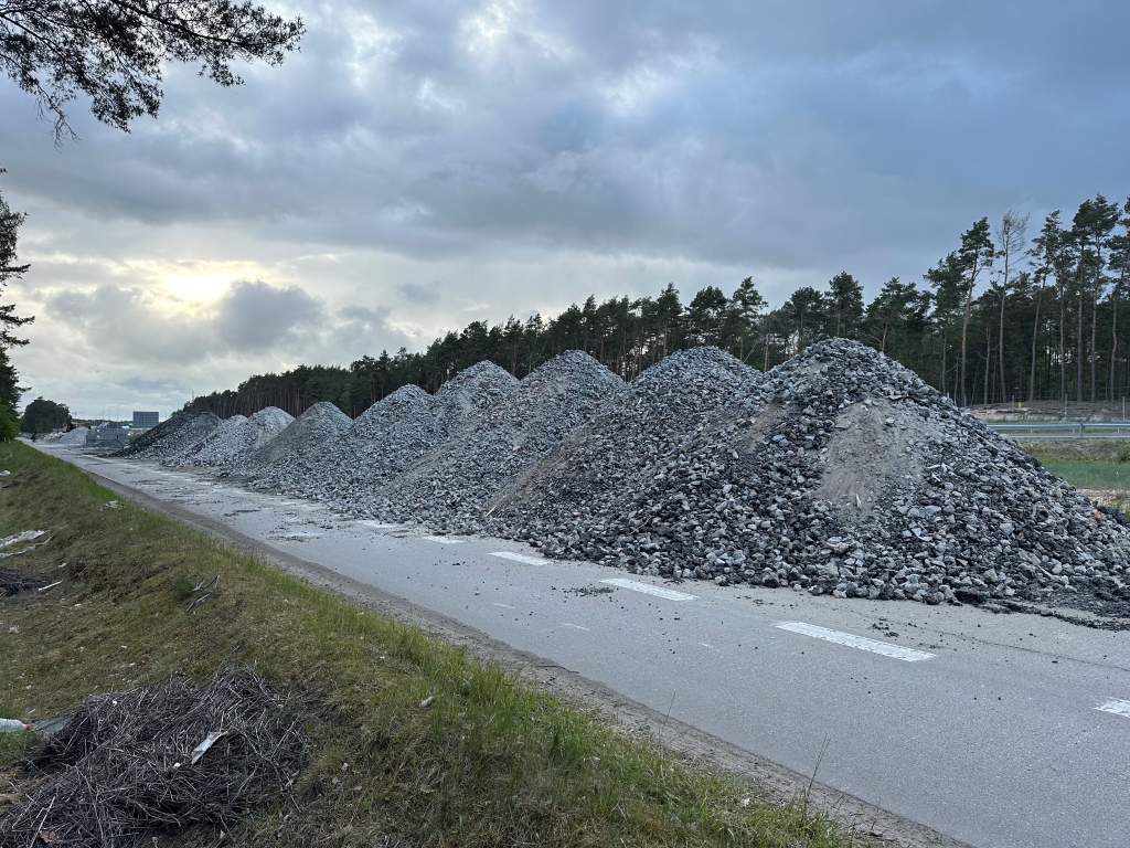 Multiple large piles of gravel lined up on a paved road beside a forest under a cloudy sky.