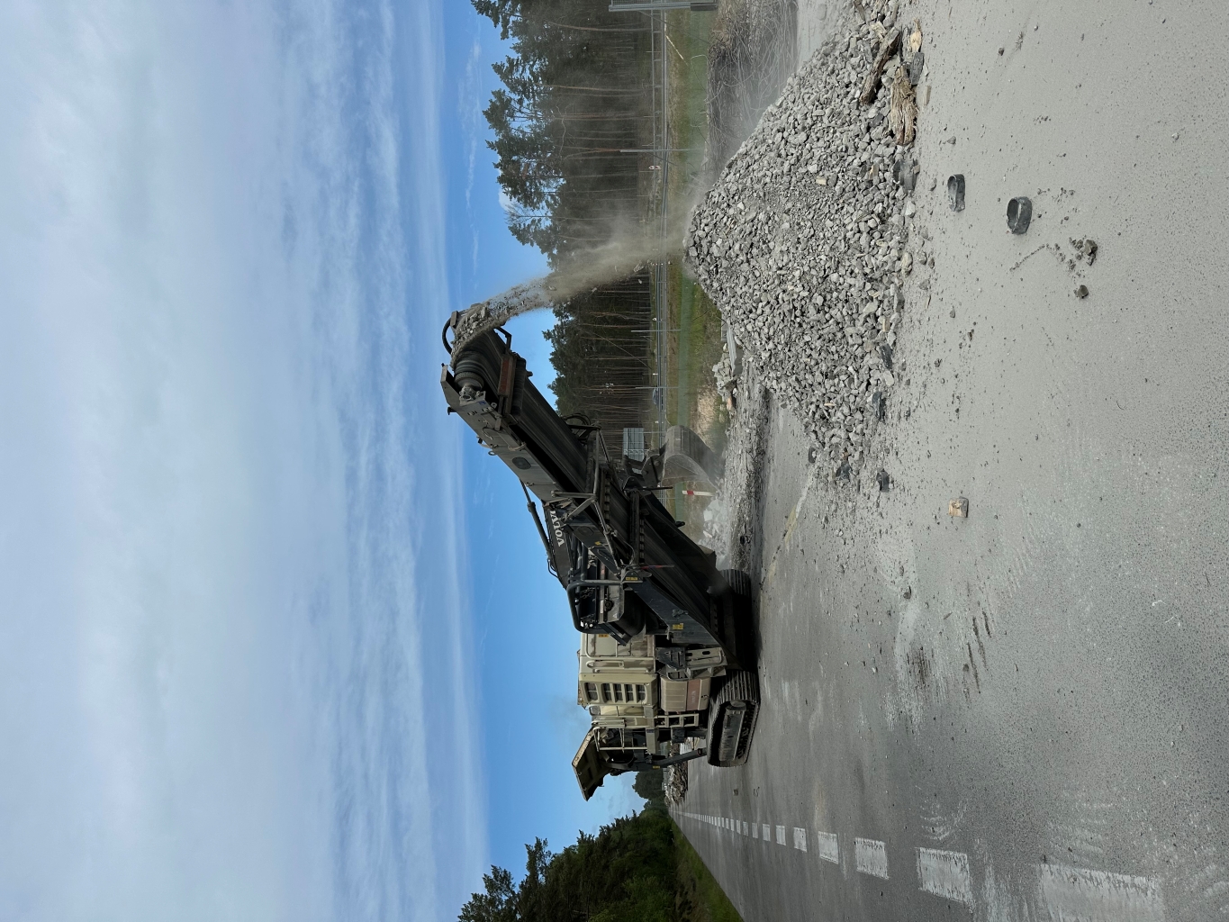 Construction machine crushing and dumping gray rocks into a pile on a road near trees under a blue sky.