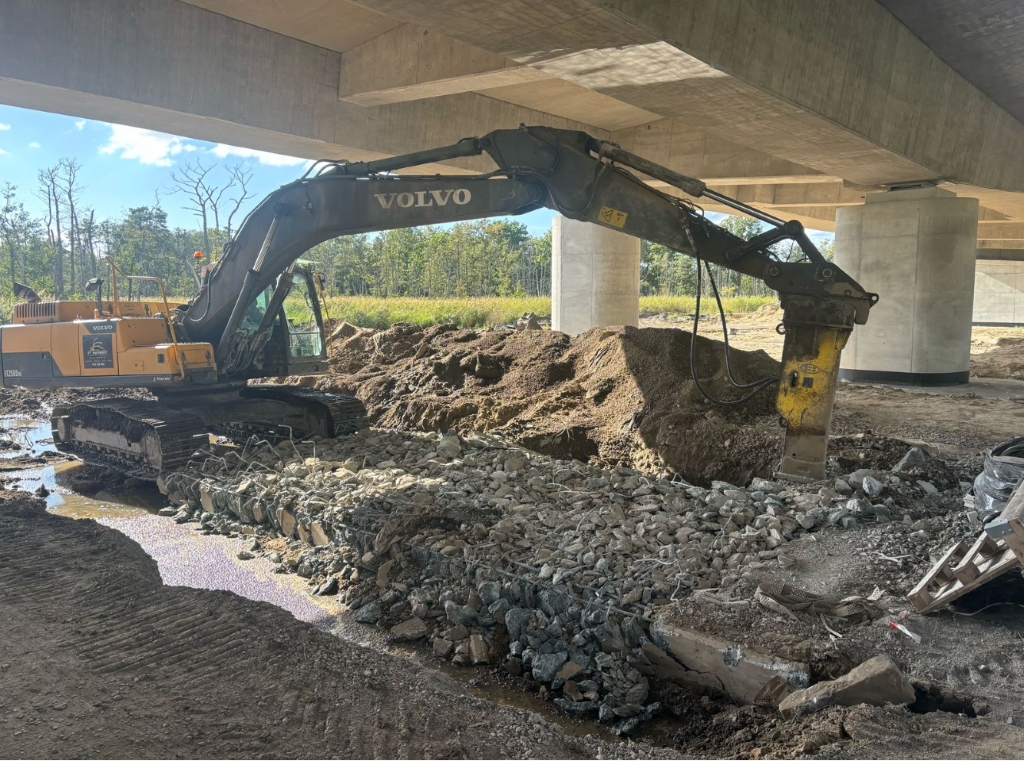 Yellow Volvo excavator with hydraulic breaker working under a concrete bridge near dirt and rocks.