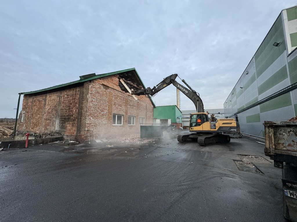 Excavator demolishing the corner of a brick building near industrial warehouses on an overcast day.