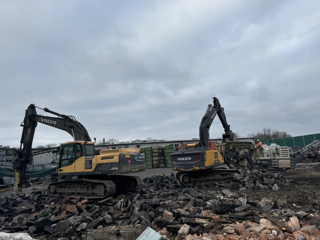Two Volvo excavators working on rubble and debris at a construction site under a cloudy sky.
