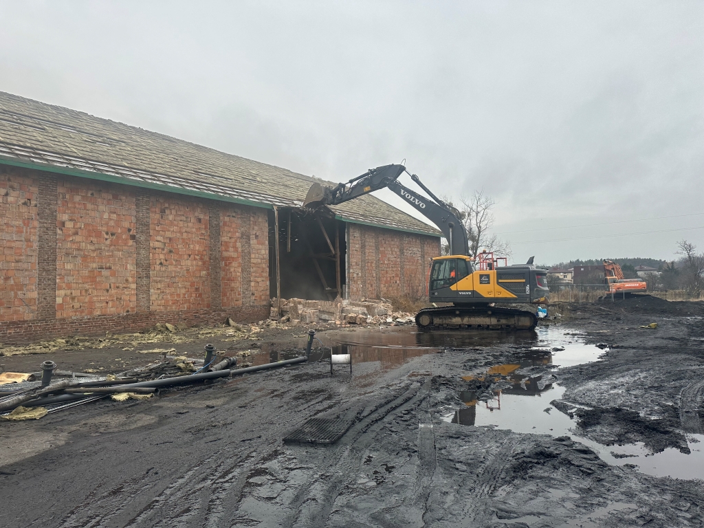 Volvo excavator demolishing a brick building on muddy ground with puddles under gray sky.