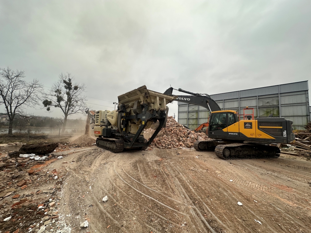 Excavator loading debris into crusher machine at a construction demolition site with piles of rubble.