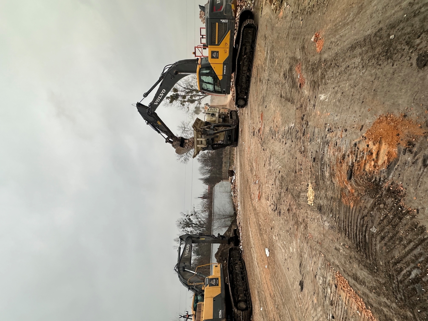 Two Volvo excavators working on a dirt site loading debris into a machine under an overcast sky.