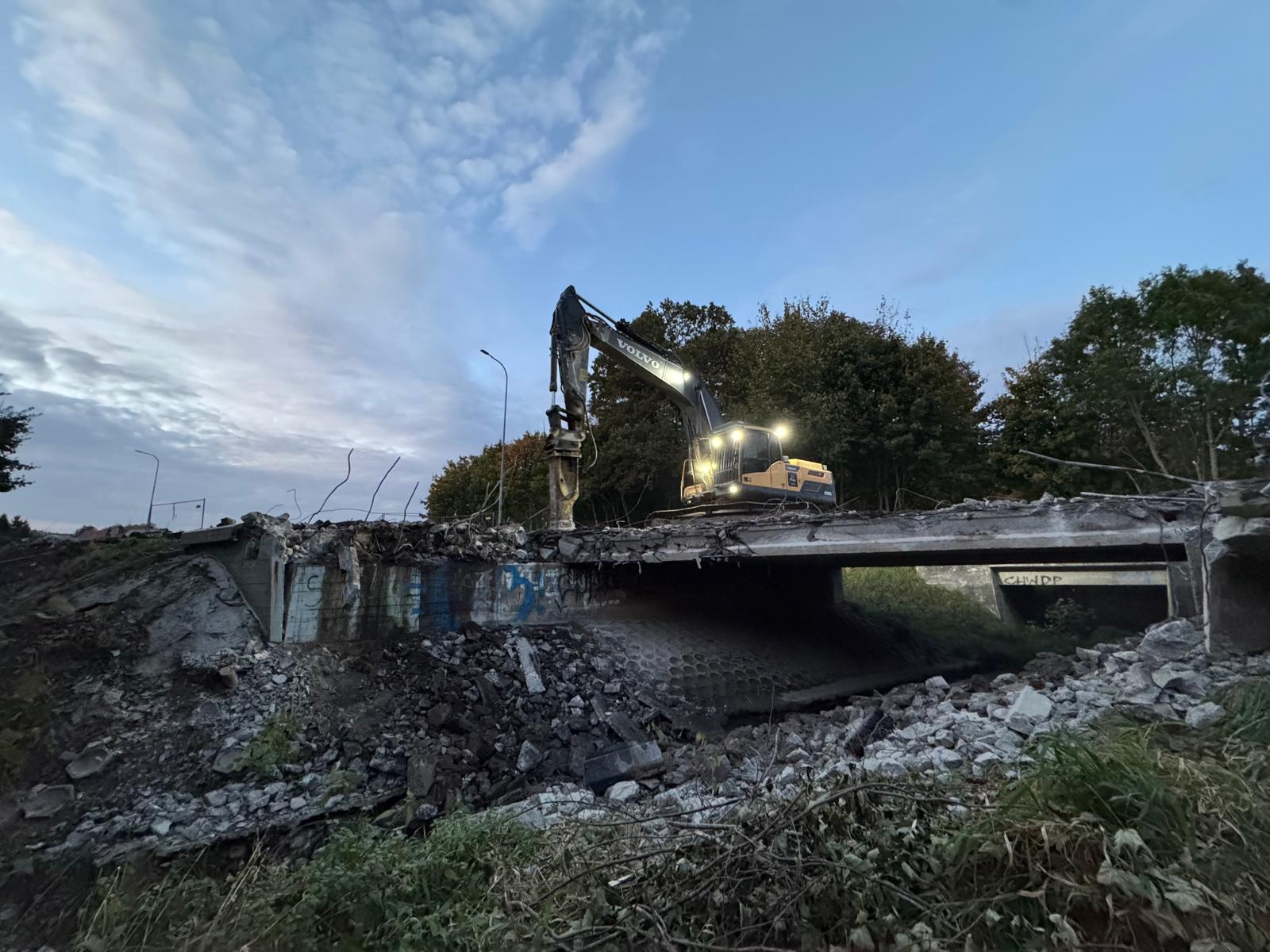 Excavator with lights on demolishing a concrete bridge surrounded by rubble and trees at dusk.