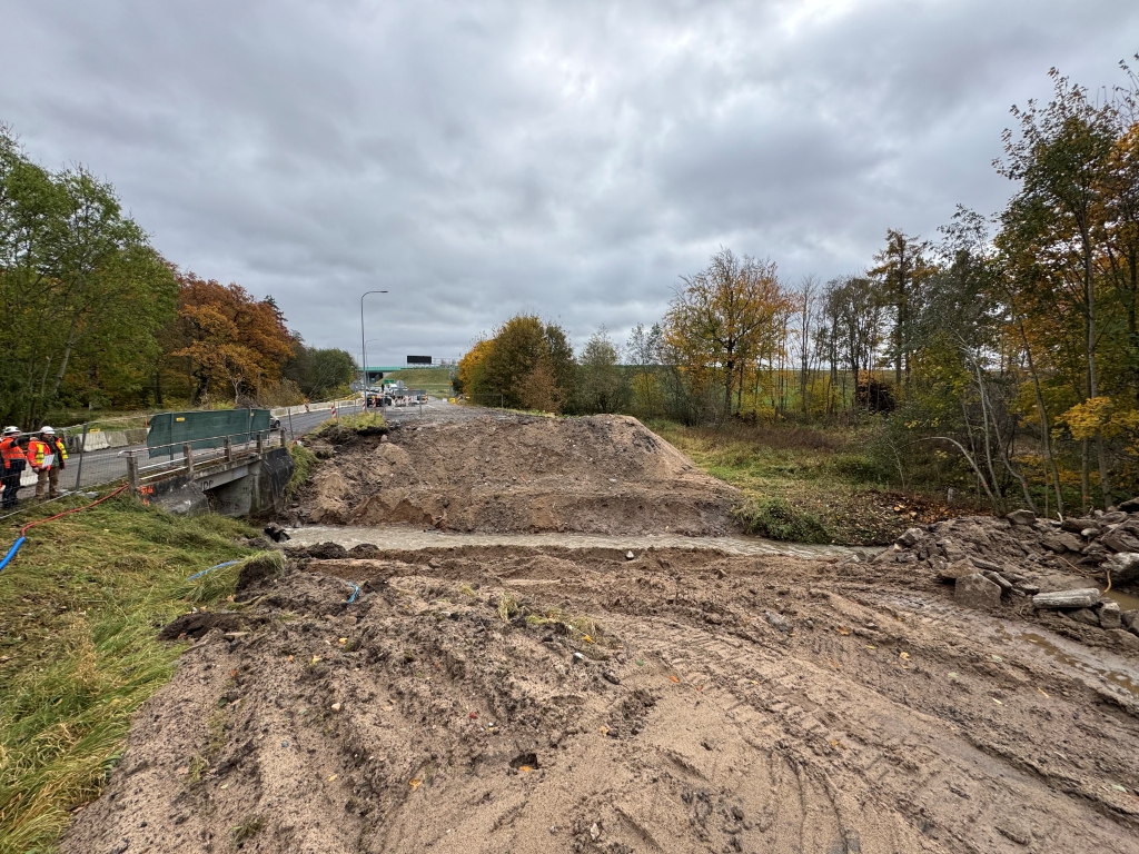 Construction site with dirt excavation near road and workers standing on left side under cloudy sky.