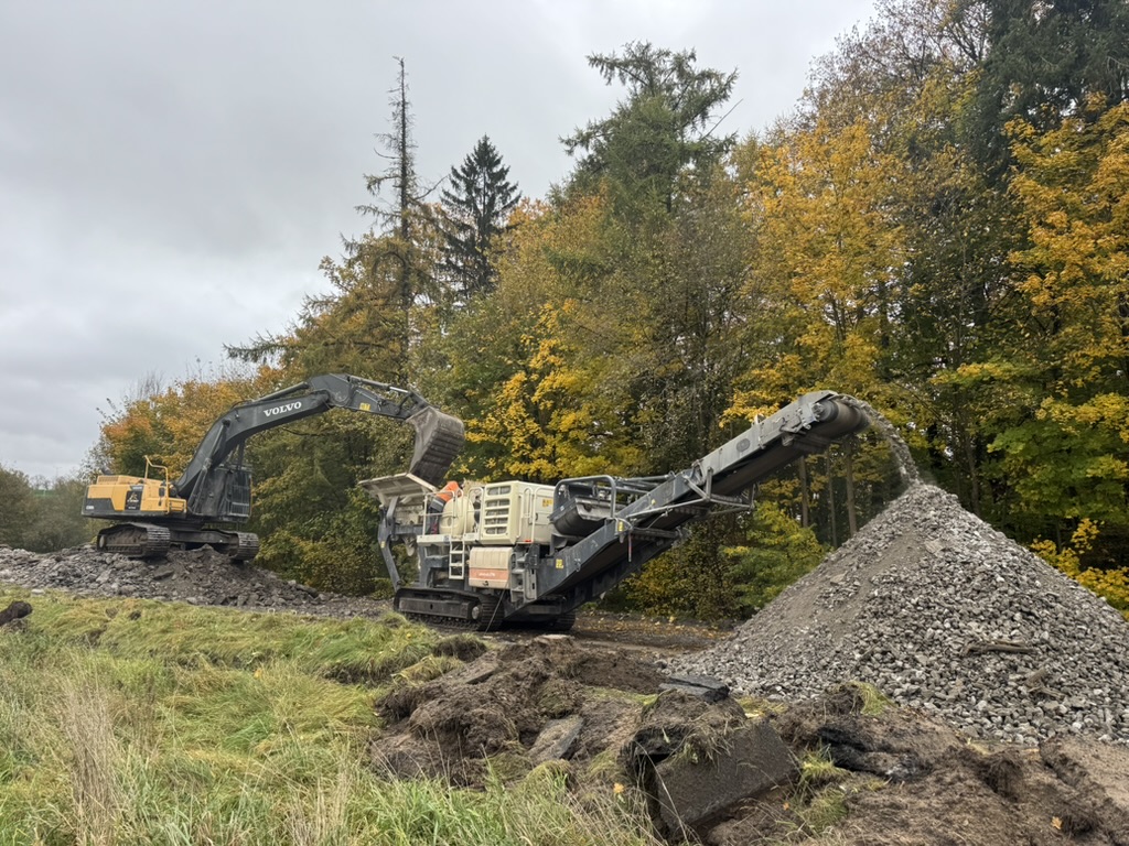 Excavator loading rocks into crusher machine that forms a pile of crushed stones outdoors.