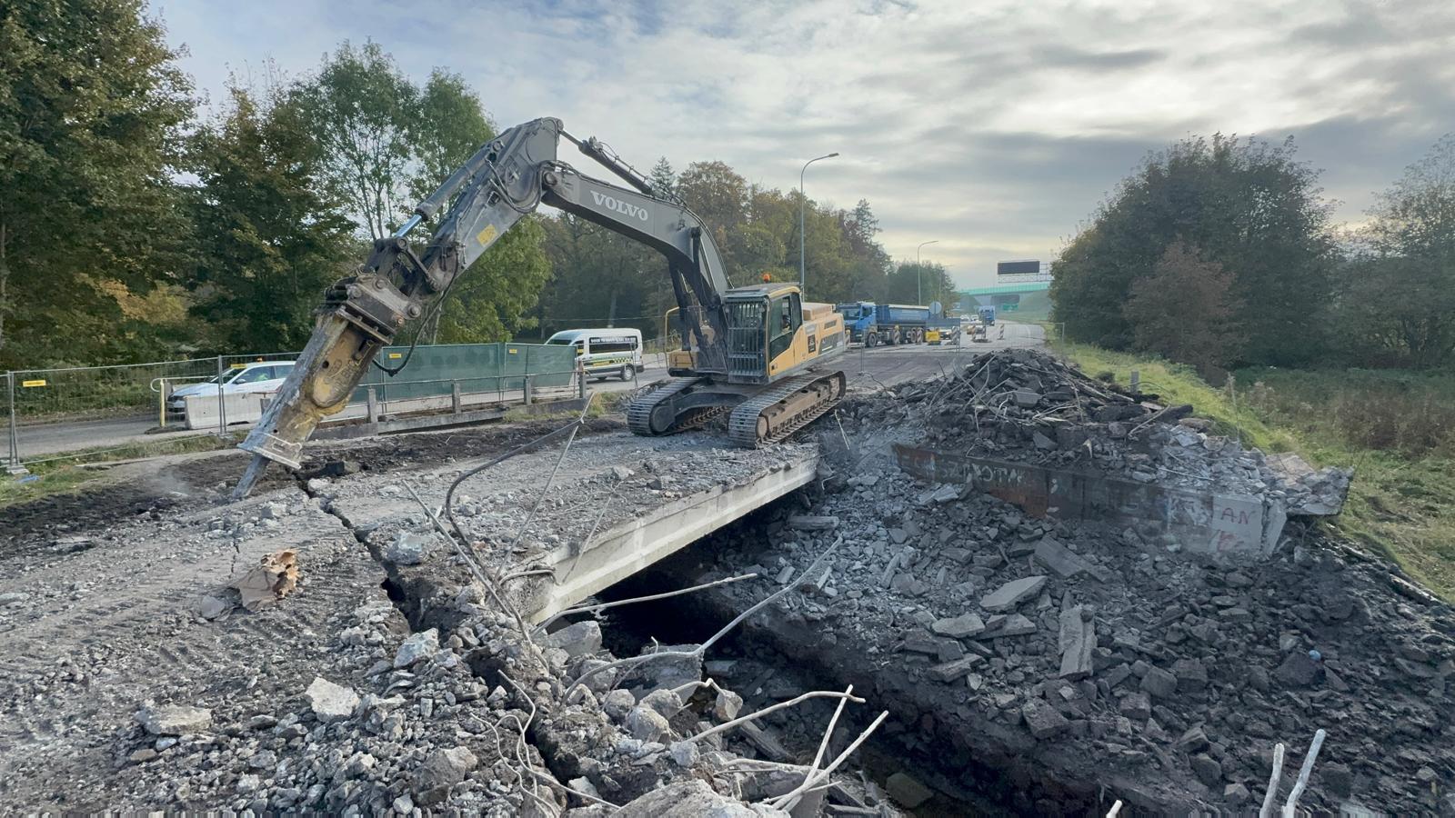Excavator demolishing a concrete bridge with rubble and exposed rebar around it on a road.