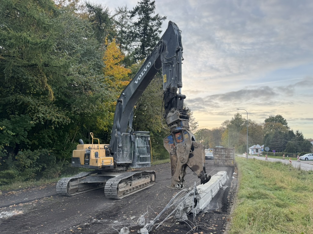 Yellow Volvo excavator demolishing concrete barriers beside a road near trees and grassy field at sunset.