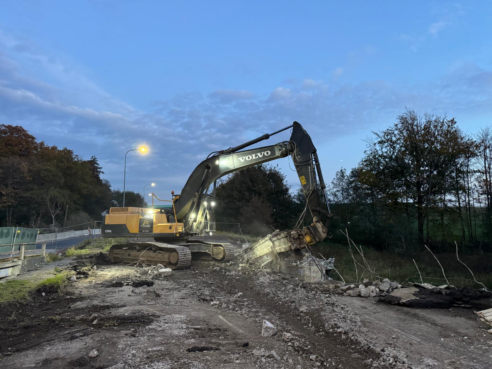 Volvo excavator demolishing concrete road section near trees at dusk with street lights on.