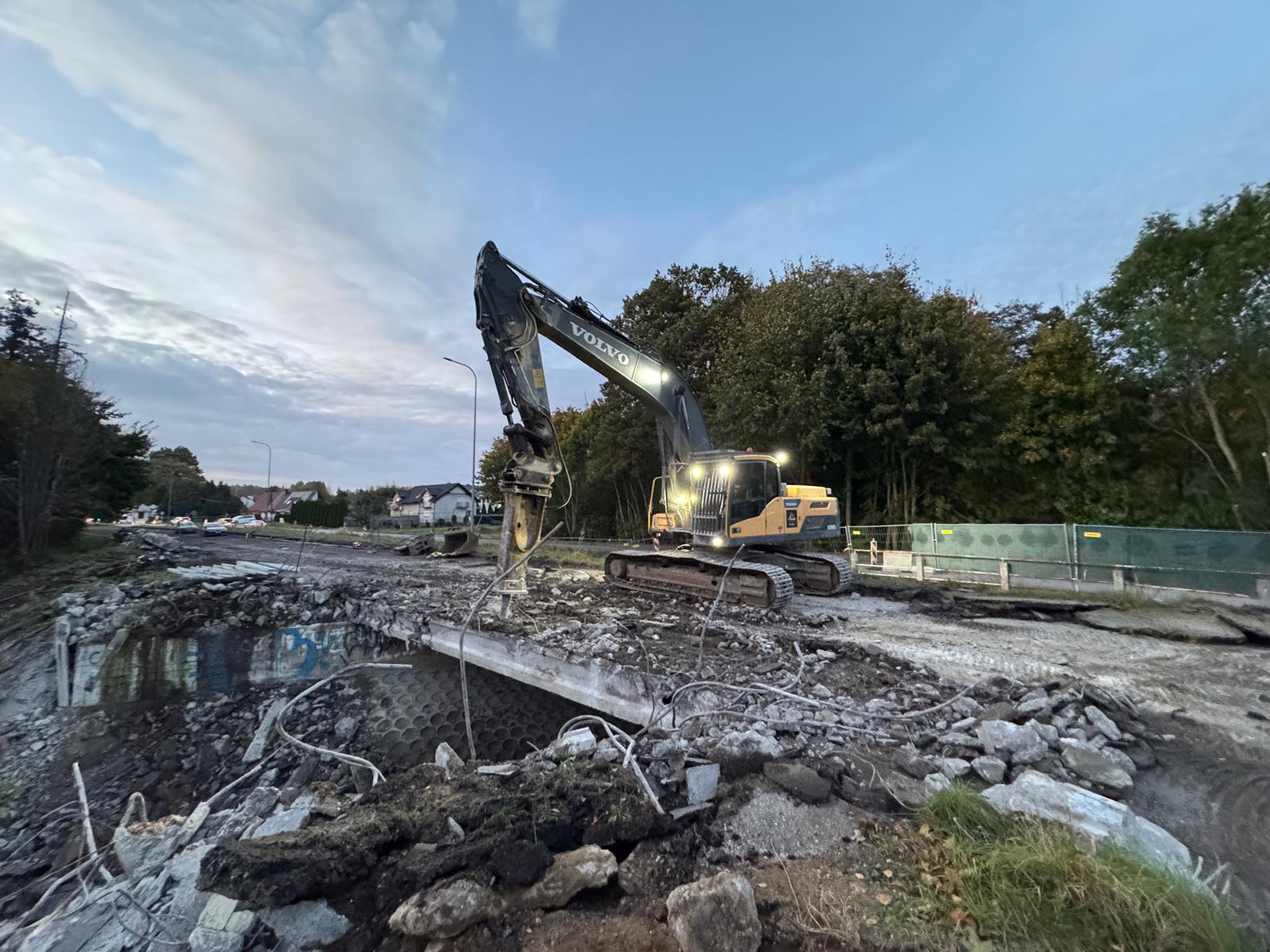 Excavator with lights demolishing a concrete bridge surrounded by rubble and trees at dusk.