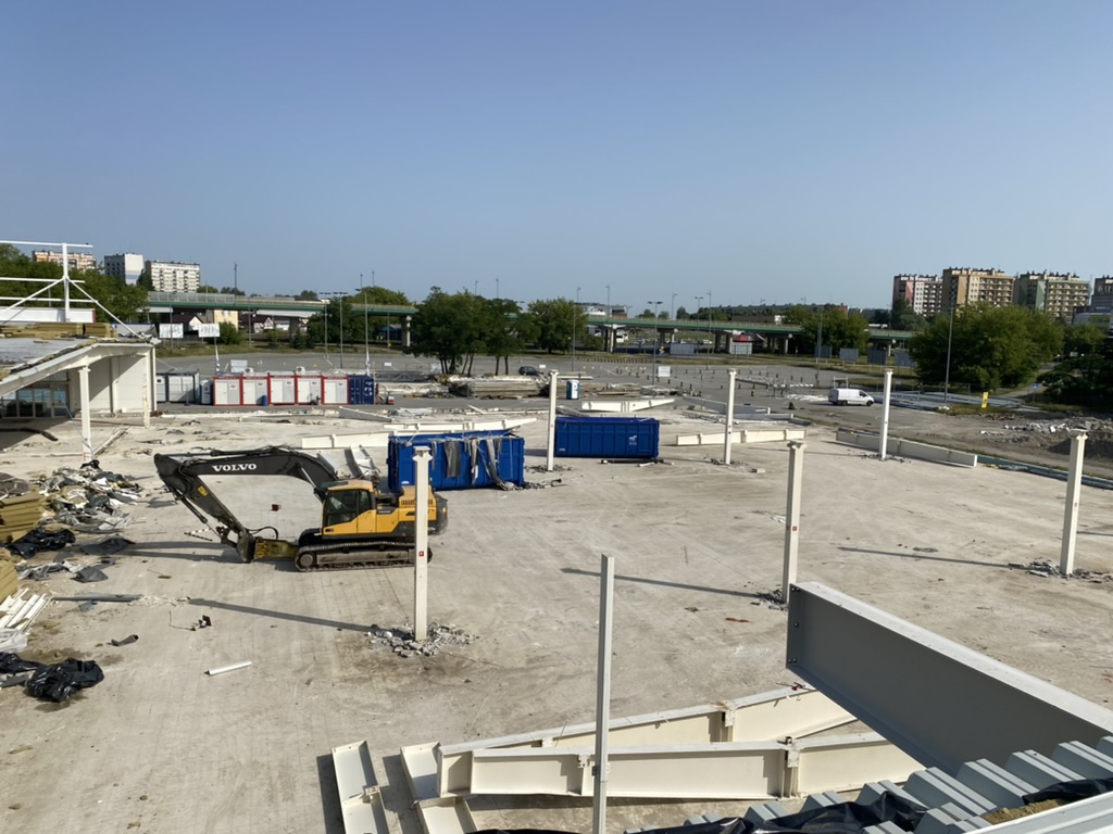 Construction site with a Volvo excavator, scattered debris, blue containers, and building columns on a clear day.
