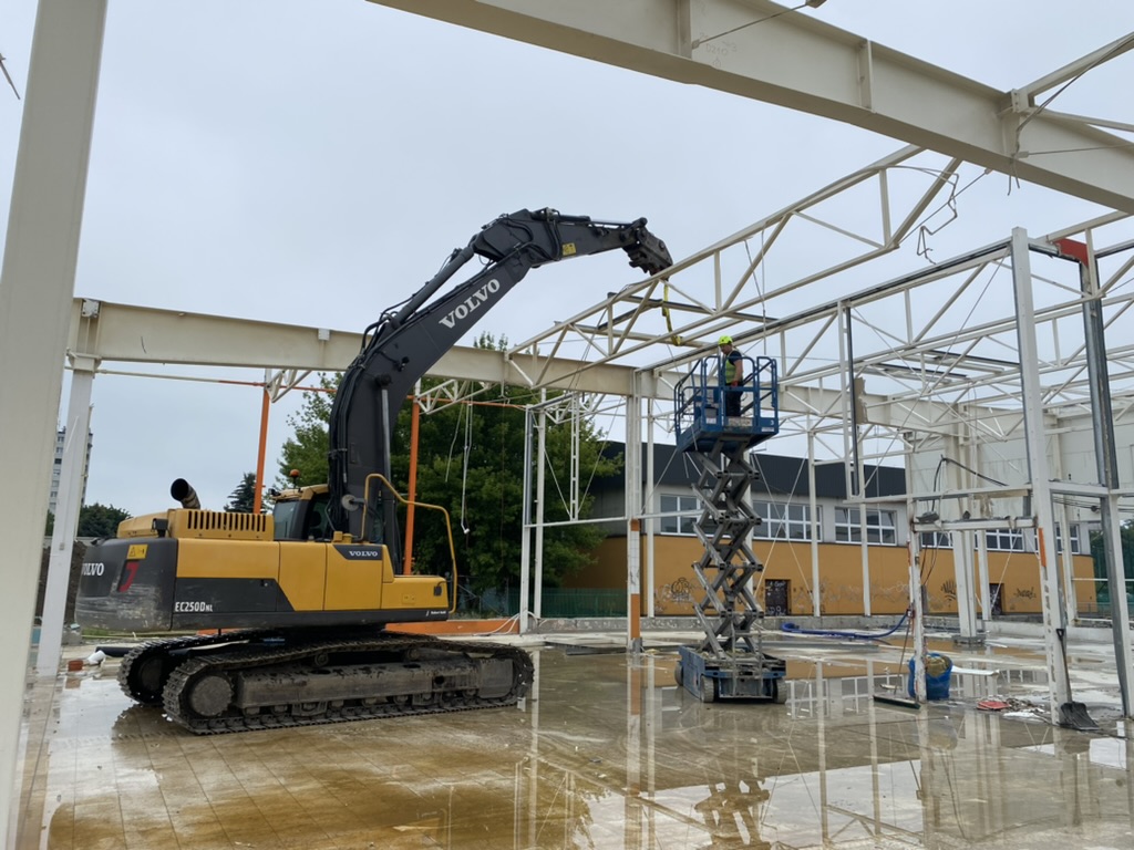 Yellow Volvo excavator and worker on a scissor lift at a construction site with steel framework.