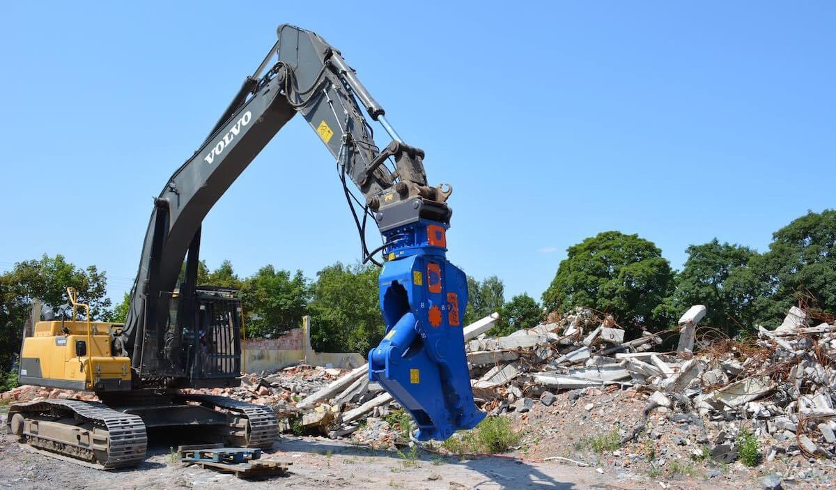 Yellow and black Volvo excavator with blue hydraulic attachment clearing construction debris outdoors.