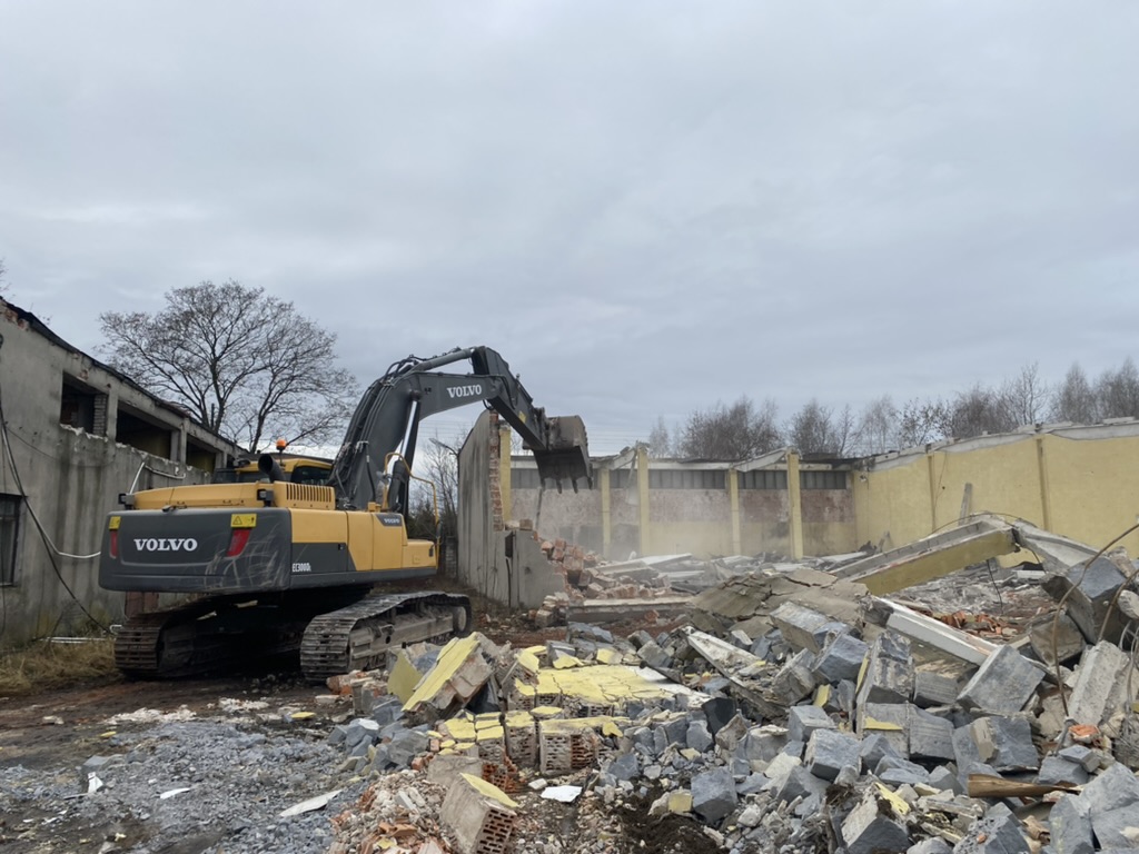 Yellow Volvo excavator demolishing a partially collapsed building with rubble and debris around it.