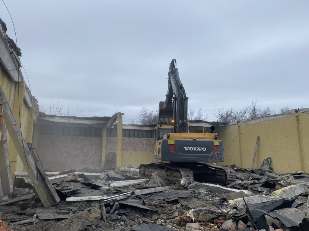 Volvo excavator demolishing debris inside a partially destroyed yellow building under cloudy sky.