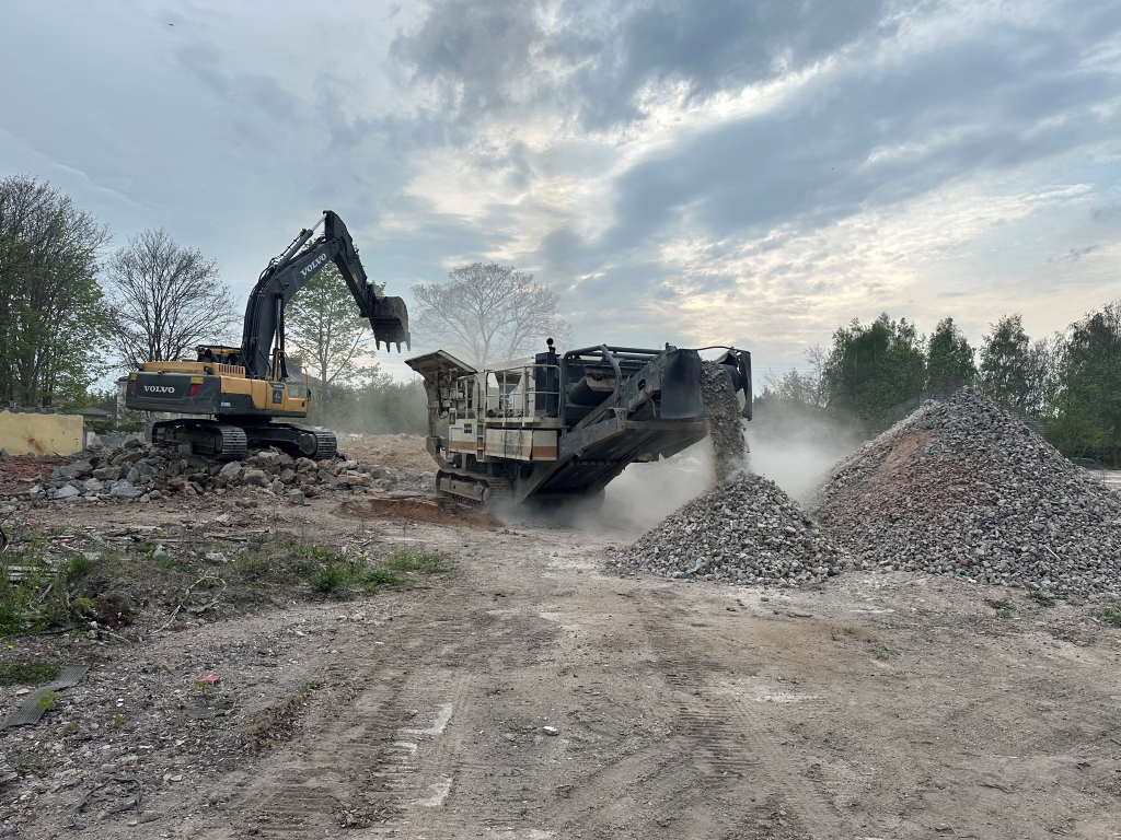Excavator loading debris into a crusher machine, creating piles of crushed rocks at a construction site.
