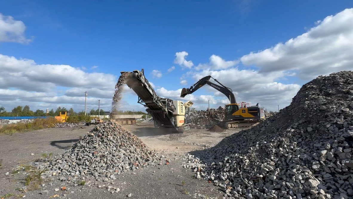 Excavator loading rubble into a crusher that deposits crushed stones in a pile under blue sky.