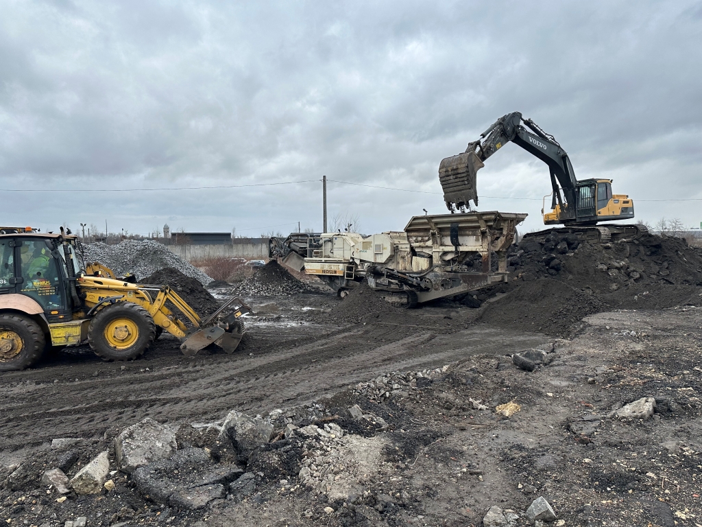 Excavator loading soil into a crusher with a loader nearby at a muddy construction site under cloudy sky.
