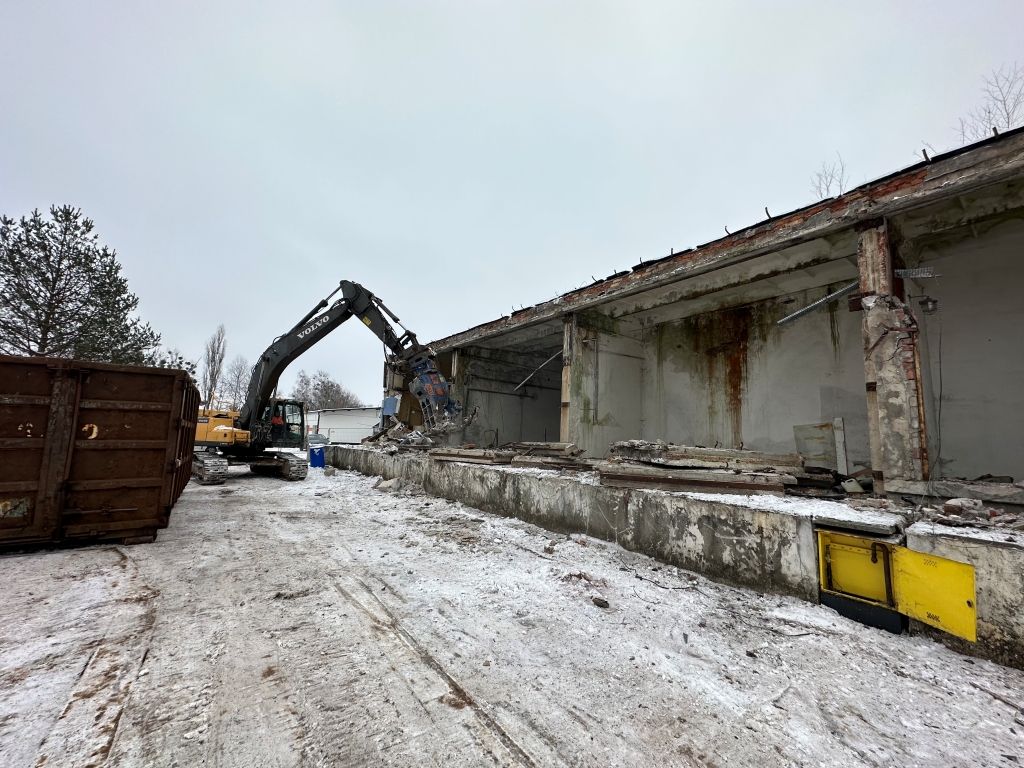 Excavator demolishing a concrete structure in a snowy industrial site under a gray sky.