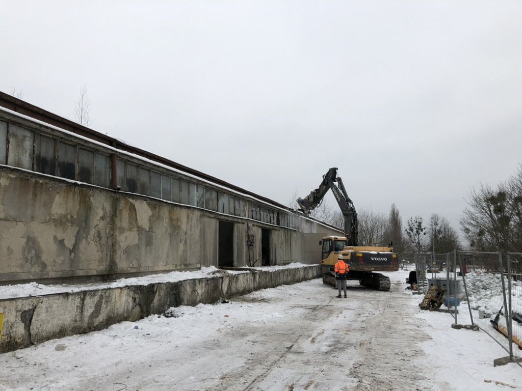 Construction worker in orange jacket standing near excavator demolishing old concrete building in snowy area.