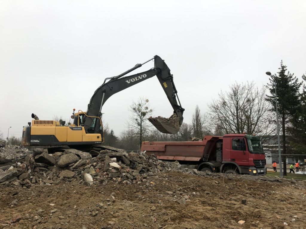 Yellow Volvo excavator loading debris into red dump truck at construction site on cloudy day.