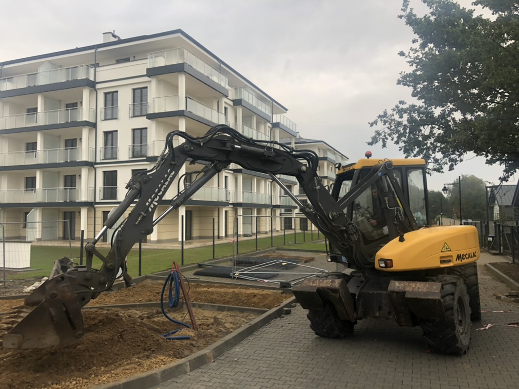 Yellow Mecalac excavator digging dirt at a construction site near a modern apartment building.