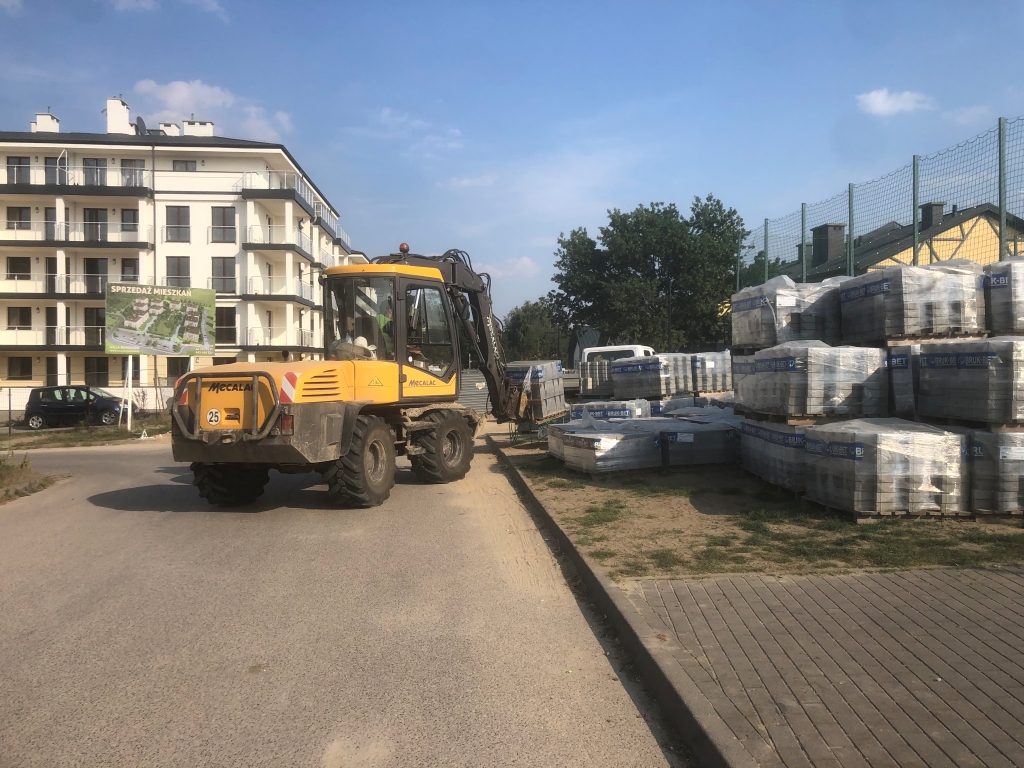 Yellow MECALAC excavator lifting pallets of bricks at a construction site near a residential building.