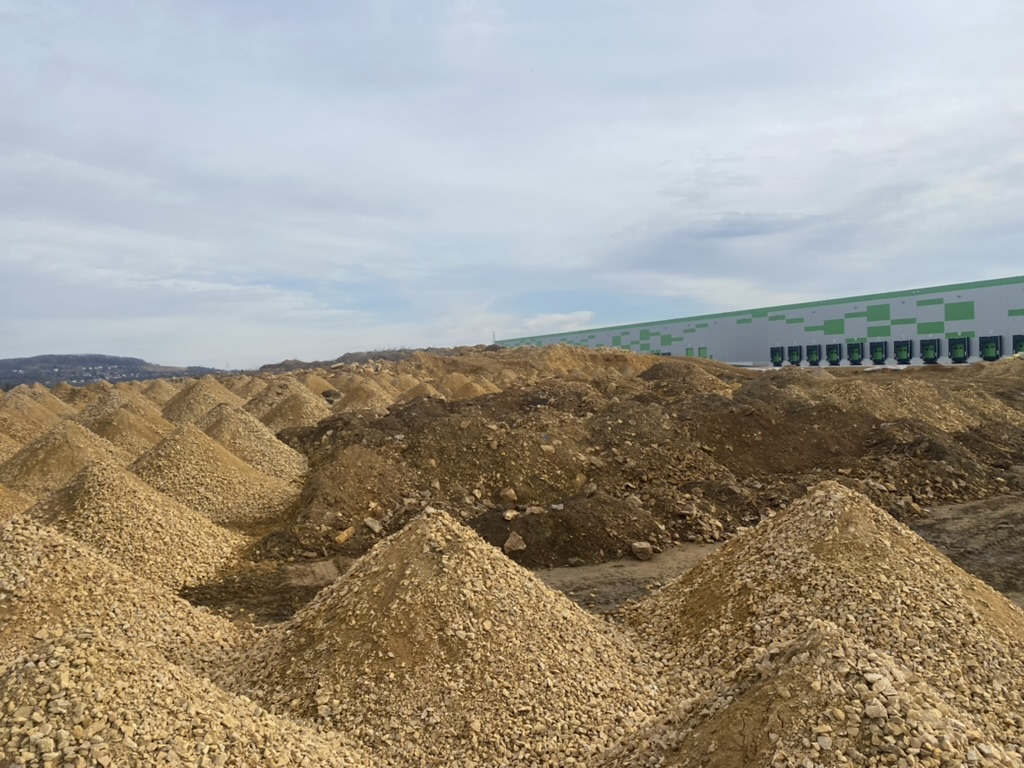 Numerous conical piles of gravel and a large building with loading docks in the background.