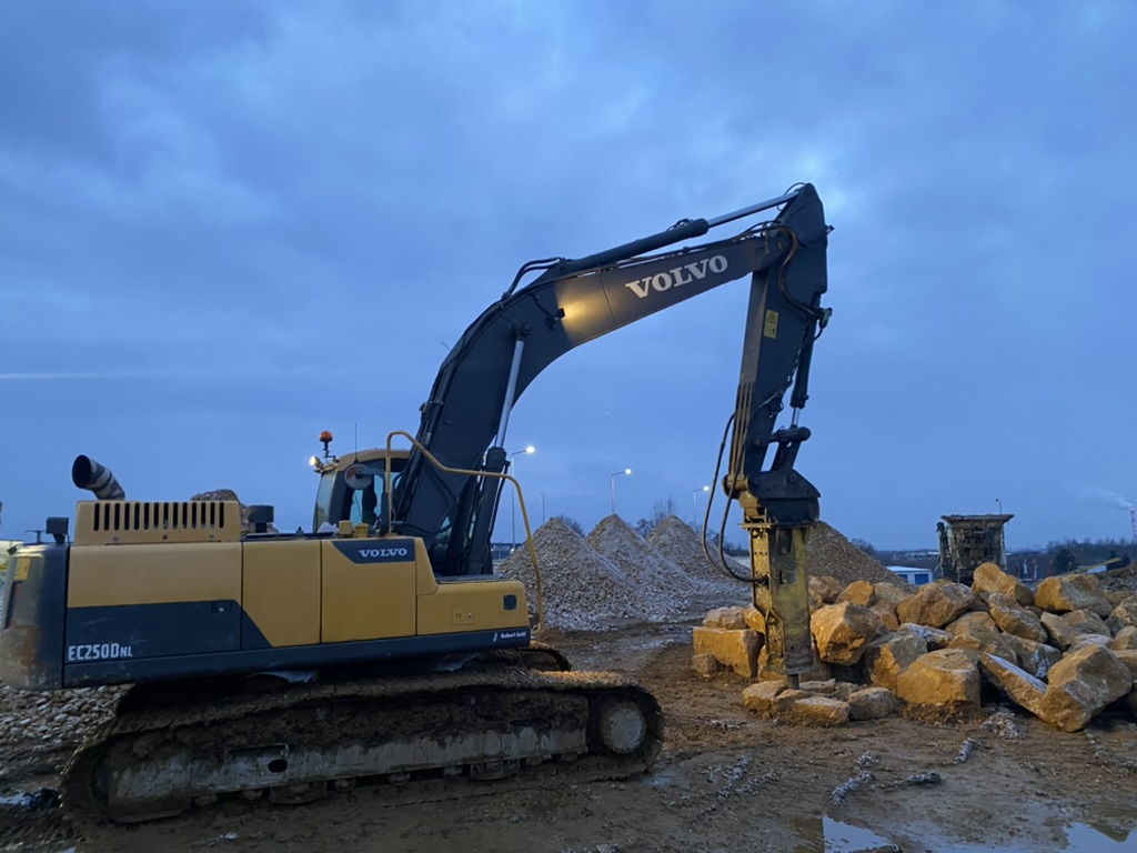 Yellow Volvo EC250DNL excavator with hydraulic hammer attachment on dirt near rock piles at dusk.