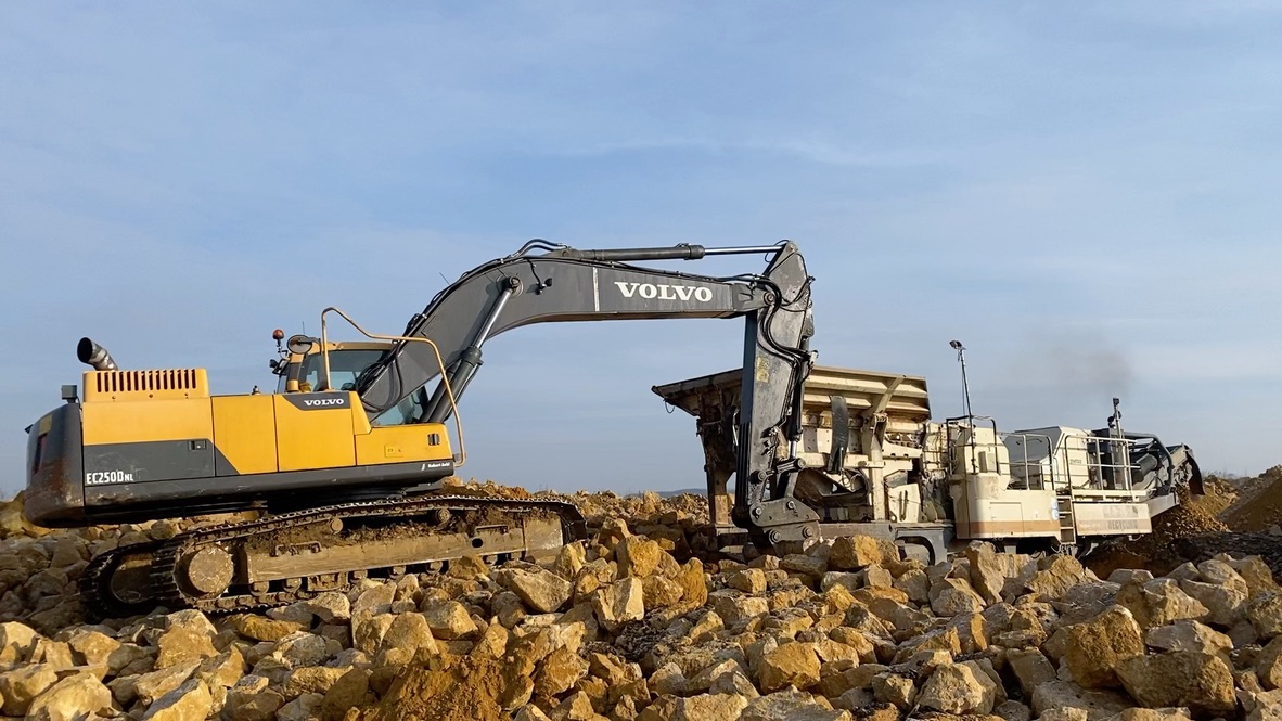 Yellow Volvo excavator with arm extended over rocky terrain against a blue sky.