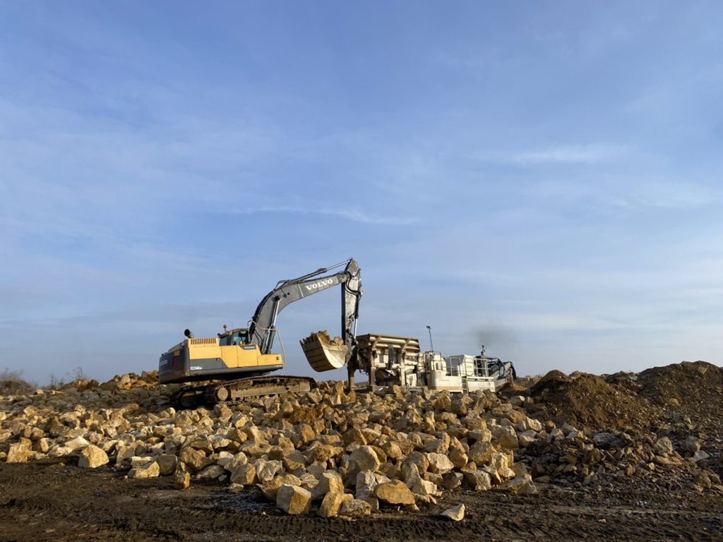Yellow Volvo excavator lifting rocks next to a rock crusher at a construction site.