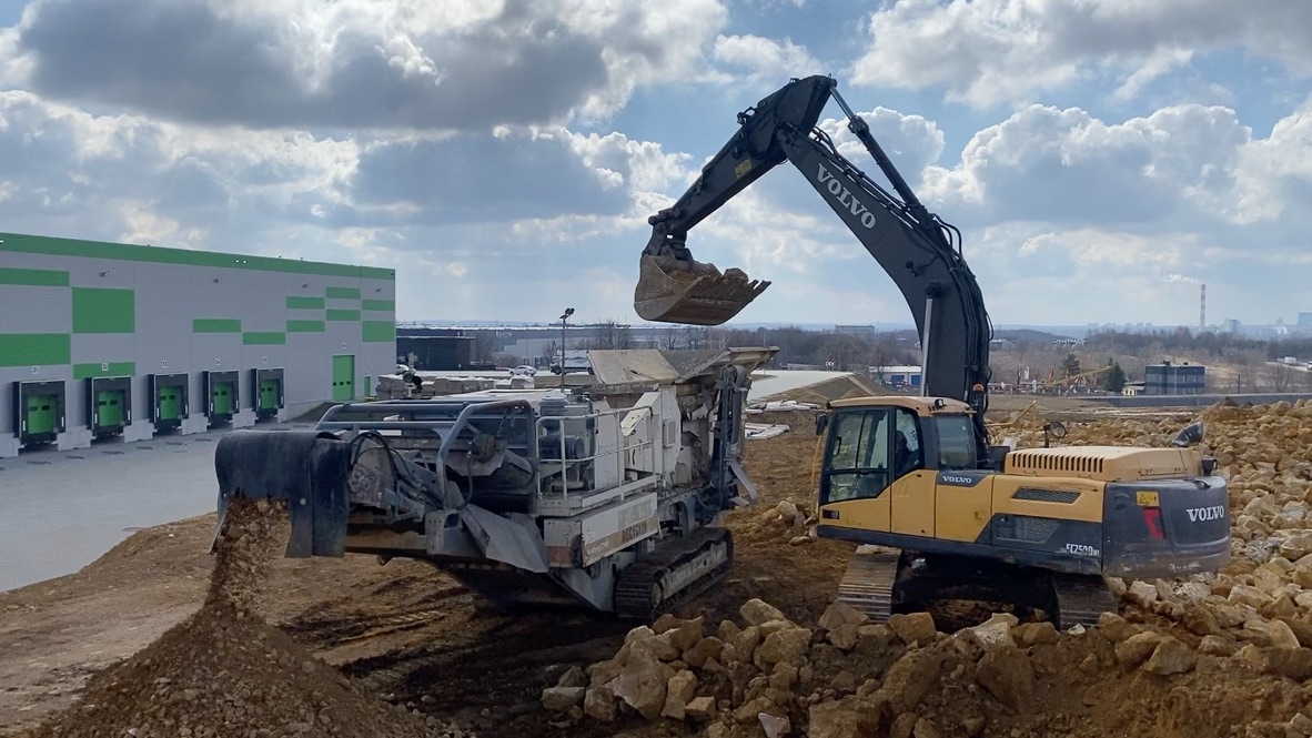 Yellow Volvo excavator dumping dirt into a crusher machine on a rocky construction site.