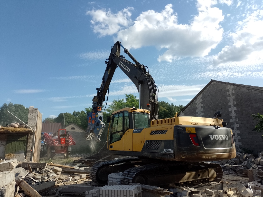 Yellow Volvo excavator with hydraulic claw amidst rubble at a demolition site under blue sky.
