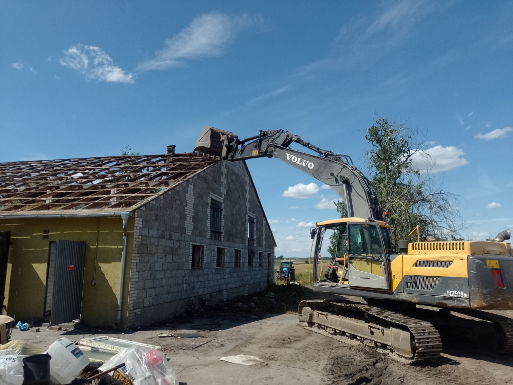 Volvo excavator demolishing the roof of a gray brick building under a blue sky.