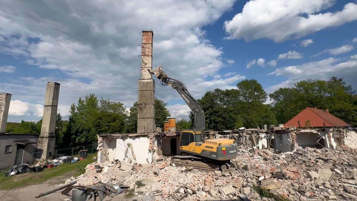 Excavator demolishing a tall chimney on a partially destroyed building under blue cloudy sky.
