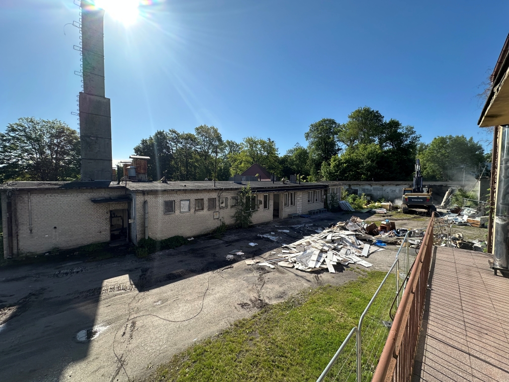 Construction area with a long building, piles of debris, an excavator, and a tall chimney under clear sky.