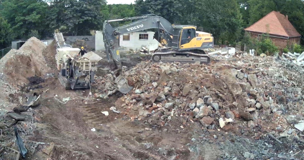 Yellow Volvo excavator and crusher machine working on rubble and dirt at a demolition site with trees and houses nearby.