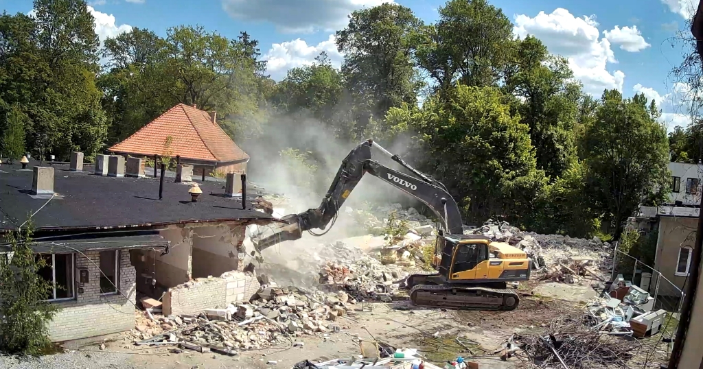 Yellow Volvo excavator demolishing a brick building amid rubble and trees under a partly cloudy sky.
