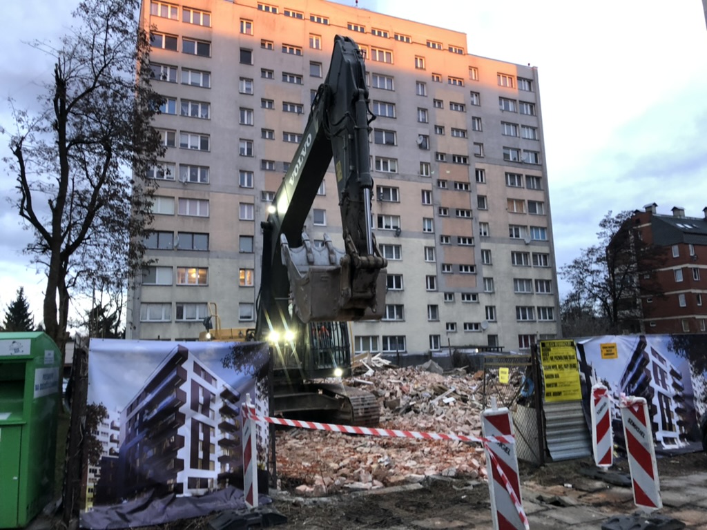 Construction site with excavator, piles of rubble, and an apartment building in the background at dusk.