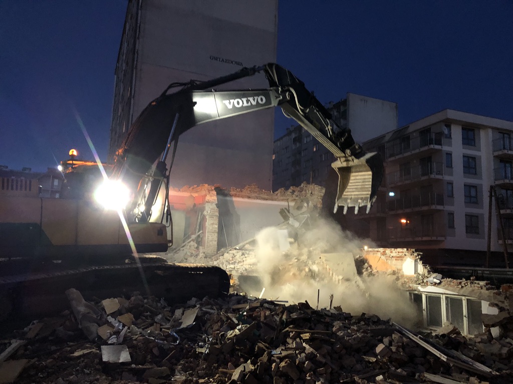 Excavator with bright lights demolishing a building at night surrounded by rubble and dust.