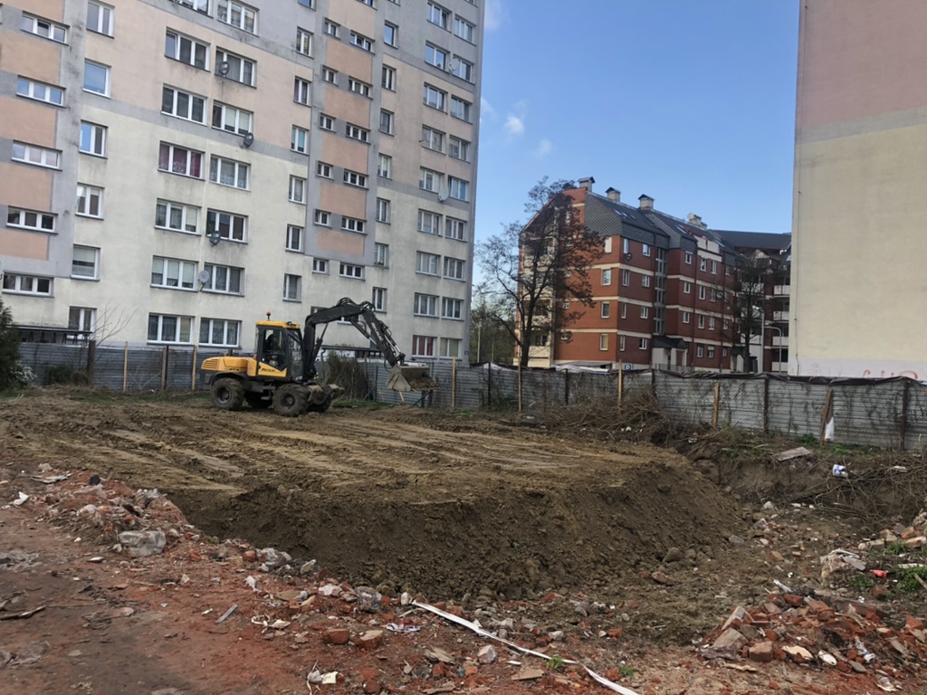 Construction site with excavator digging dirt next to tall apartment buildings and a clear sky.