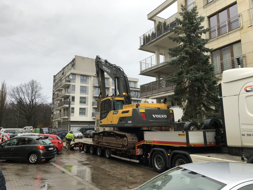 Yellow Volvo excavator on flatbed truck trailer parked on wet street near buildings and cars.