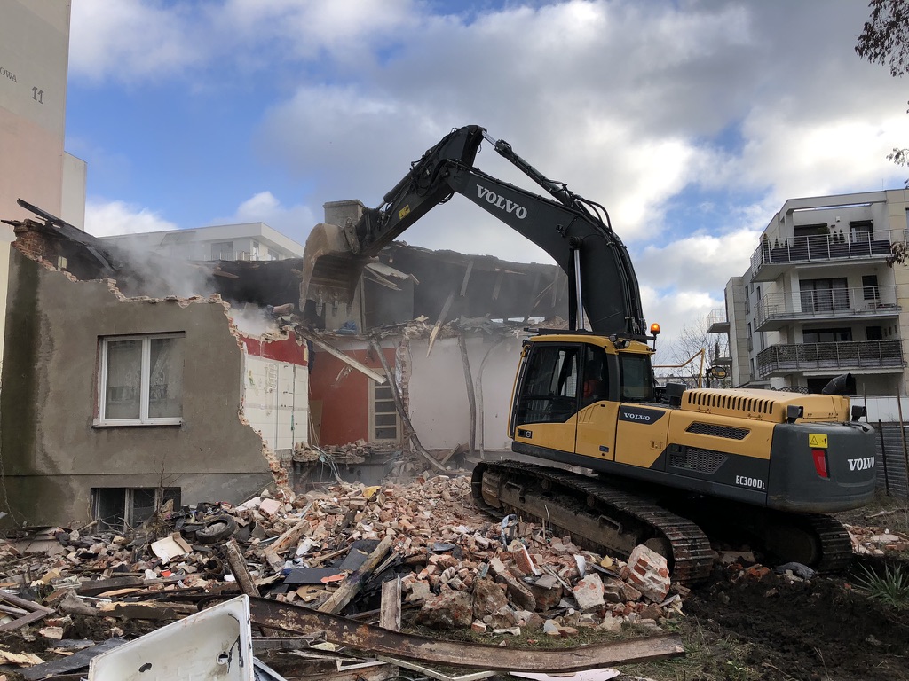 Yellow Volvo excavator demolishing a partially destroyed brick building amid rubble.