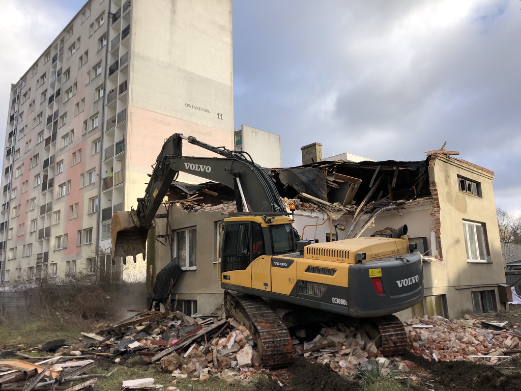 Yellow Volvo excavator demolishes a partially destroyed two-story building near an apartment block.