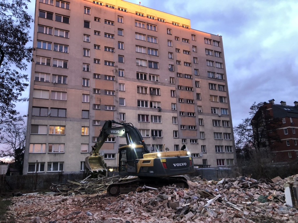 Volvo excavator with lights on clearing rubble in front of a tall apartment building at dusk.