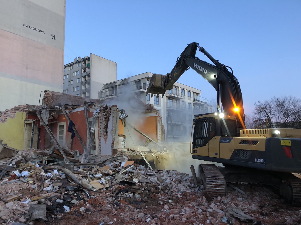 Excavator demolishing a brick building amid rubble at dusk with residential buildings in background.